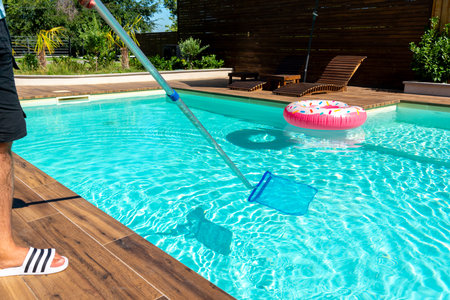 Man cleaning turquoise swimming pool water with skimmer net, wooden deck, and colorful inflatable floatsの写真素材