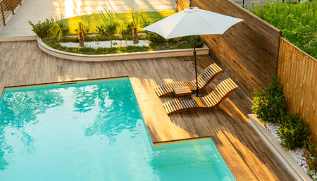 Luxurious poolside wooden deck with curved lounge chairs, umbrella, and turquoise swimming pool in a sunny backyardの写真素材