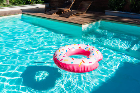 Pink donut-shaped inflatable float in turquoise swimming pool with wooden deck and sunny reflectionsの写真素材