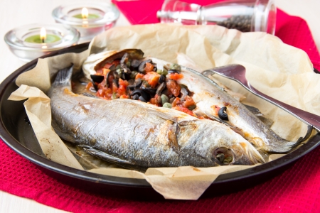 Baked whole white fish, sea bass stuffed with black olives, capers and tomatoesの写真素材