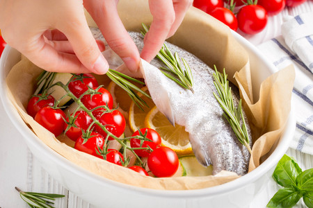 Cooking dinner, raw white trout fish in a baking dish with zucchini, cherry tomatoes on a branch, rosemary and lemon. Delicious healthy diet foodの写真素材