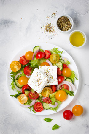 Tasty greek salad with colorful cherry tomatoes red and yellow, cucumber, onion, lettuce and large piece of feta cheese with herbs. In white plate on light backgroundの写真素材