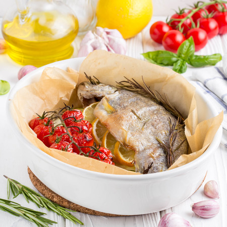 Baked whole white trout fish in ceramic baking dish with zucchini, cherry tomatoes on branch, rosemary and lemon. Delicious healthy diet foodの写真素材