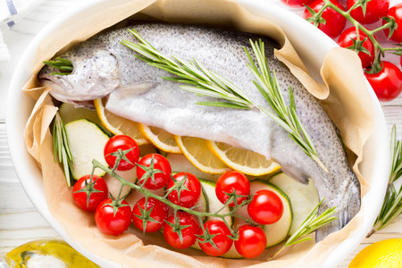 Cooking dinner, raw white trout fish in a baking dish with zucchini, cherry tomatoes on a branch, rosemary and lemon. Delicious healthy diet foodの写真素材
