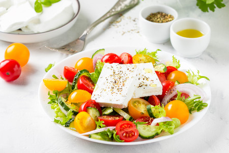 Tasty greek salad with colorful cherry tomatoes red and yellow, cucumber, onion, lettuce and large piece of feta cheese with herbs. In white plate on light backgroundの写真素材