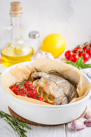 Baked whole white trout fish in ceramic baking dish with zucchini, cherry tomatoes on branch, rosemary and lemon. Delicious healthy diet foodの写真素材