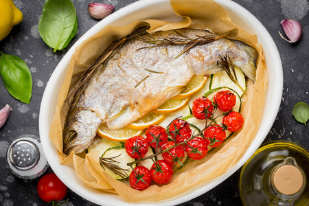 Baked whole white trout fish in ceramic baking dish with zucchini, cherry tomatoes on branch, rosemary and lemon. Delicious healthy diet foodの写真素材