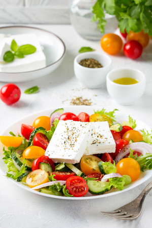 Tasty greek salad with colorful cherry tomatoes red and yellow, cucumber, onion, lettuce and large piece of feta cheese with herbs. In white plate on light backgroundの写真素材