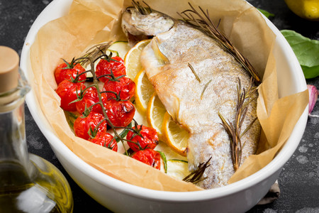 Baked whole white trout fish in ceramic baking dish with zucchini, cherry tomatoes on branch, rosemary and lemon. Delicious healthy diet foodの写真素材