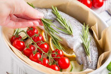 Cooking dinner, raw white trout fish in a baking dish with zucchini, cherry tomatoes on a branch, rosemary and lemon. Delicious healthy diet foodの写真素材