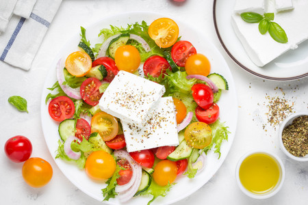 Tasty greek salad with colorful cherry tomatoes red and yellow, cucumber, onion, lettuce and large piece of feta cheese with herbs. In white plate on light backgroundの写真素材