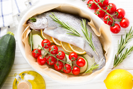 Cooking dinner, raw white trout fish in a baking dish with zucchini, cherry tomatoes on a branch, rosemary and lemon. Delicious healthy diet foodの写真素材