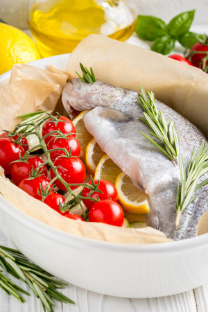 Cooking dinner, raw white trout fish in a baking dish with zucchini, cherry tomatoes on a branch, rosemary and lemon. Delicious healthy diet foodの写真素材
