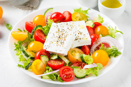 Tasty greek salad with colorful cherry tomatoes red and yellow, cucumber, onion, lettuce and large piece of feta cheese with herbs. In white plate on light backgroundの写真素材