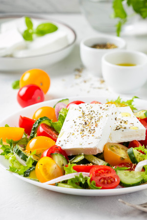 Tasty greek salad with colorful cherry tomatoes red and yellow, cucumber, onion, lettuce and large piece of feta cheese with herbs. In white plate on light backgroundの写真素材