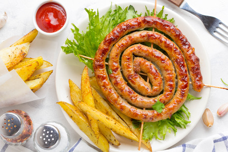 Grilled sausages with herb potatoes and ketchup, delicious summer picnic dish, sausages on skewers, twisted in a circle on a light backgroundの写真素材