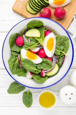 Fresh spring salad with spinach, radish, cucumber and egg. Delicious lunch, healthy food, summer vegetables. In bowl on white wooden backgroundの写真素材