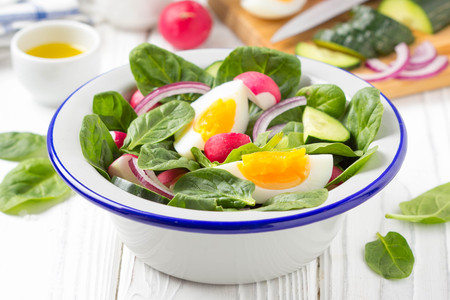 Fresh spring salad with spinach, radish, cucumber and egg. Delicious lunch, healthy food, summer vegetables. In bowl on white wooden backgroundの写真素材