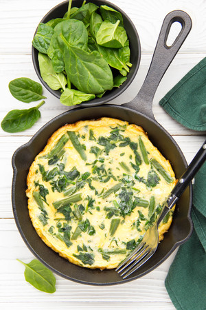Omelet with spinach and green beans, healthy food. Egg and milk Frittata, delicious Breakfast. In black frying pan on white wooden backgroundの写真素材