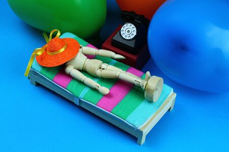 Wooden mannequin with orange straw hat lying on a beach bed. Beside old black telephone from the 50s. Shot from above, light blue background with colored balloons.の写真素材