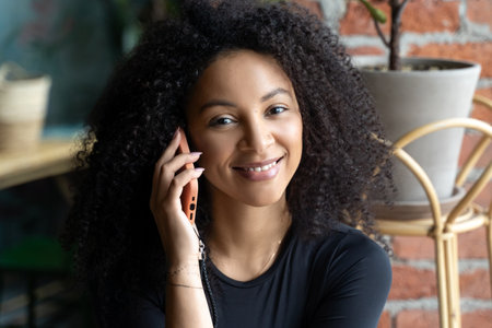 Black happy woman with African curls speaking on cellphone at home or cafe indoors. Communication with friends of relatives. Comfort relationships. Freelanceの写真素材