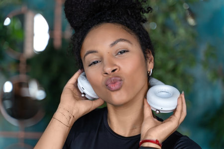 Beautiful young black woman wearing stylish white headphones on dark loft apartment background. Copy...