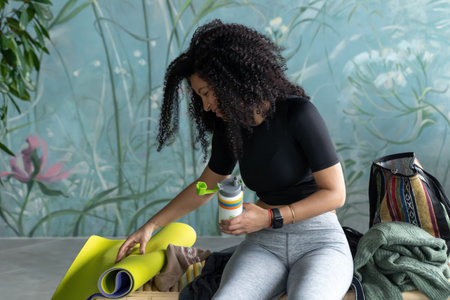 Middle-aged multiethnic woman packing sport stuff and accessories after fitness training over green domestic house plant loft blue home background. lifestyle concept. Copy space, mock-up. Eco natural, body consciousの写真素材