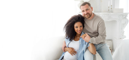 Caucasian man embrace his pregnant black wife. Pregnant woman and loving husband hugging tummy at home, smile at camera. Bannerの写真素材