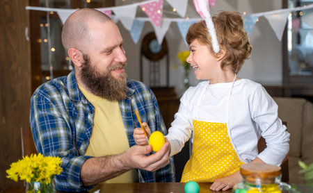 Easter Family traditions. Father and caucasian happy child with bunny ears dye and decorate eggs with paints for holidays while sitting together at home table having funの写真素材