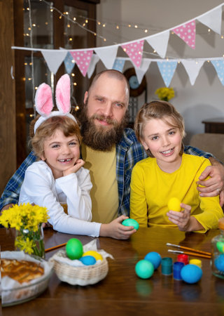 Easter Family traditions. Father and two caucasian happy children with bunny ears dye and decorate eggs with paints for holidays while sitting together at home table. Kids embrace and smile in cozy.の写真素材