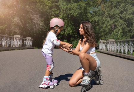 Mother teaching child daughter to skate on inline skates rollers in public park in summer. Family...
