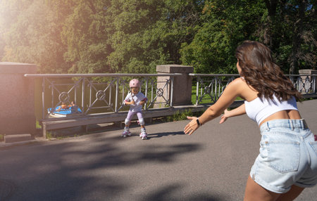 Mother teaching child daughter to skate on inline skates rollers in public park in summer. Family leisure outdoor sport activity game. Copy negative spaceの写真素材
