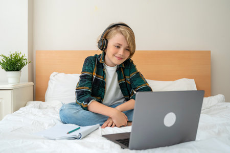 Teenage child boy using laptop and headphones. Child looking online video course lesson in bed. Chat video conference Schoolboy studying doing homework. E-learning, remote educational classesの写真素材
