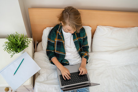 Teenage child boy using laptop. Child typing on the keyboard lying in bed. Play video game. Schoolboy studying doing homework. E-learning, remote educational classes top viewの写真素材