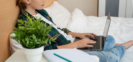 Teenage child boy using laptop. Child typing on the keyboard lying in bed. Play video game. Schoolboy studying doing homework. E-learning, remote educational classesの写真素材