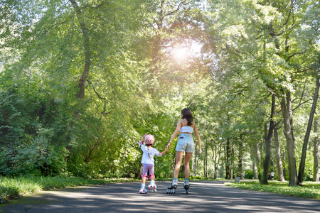 Mother teaching child daughter to skate on inline skates rollers in public park in summer. Family leisure outdoor sport activity game. Copy negative spaceの写真素材