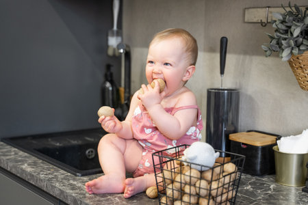 Little caucasian baby girl sitting on table in domestic kitchen. Cute toddler wear bodysuit and play with nuts. Daily home routine. Goods and products for children advertisement copy spaceの写真素材