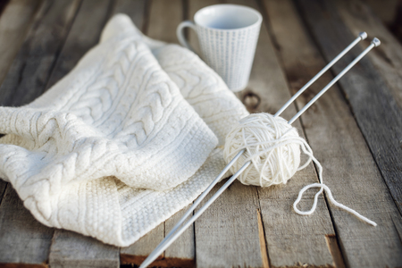 Beautiful white knitted scarf and ball of wool with needles and cup of tea on old wooden table. Cozy...