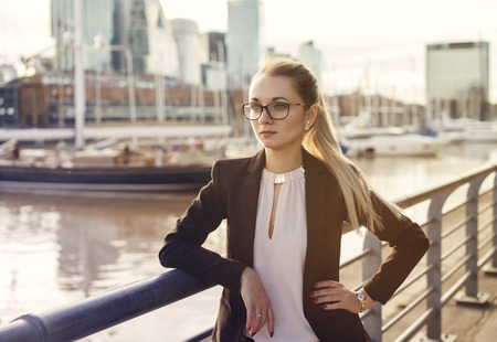 Blonde business woman in suit with glasses standing near the river on city backgroundの写真素材