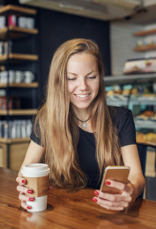 Beautiful young woman writing a message in the cell phone sitting in the cafe with a cup of coffee in her handの写真素材
