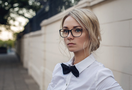 Portrait of beautiful blonde girl in glasses, white shirt and black bow tie outdoors, copy space,...