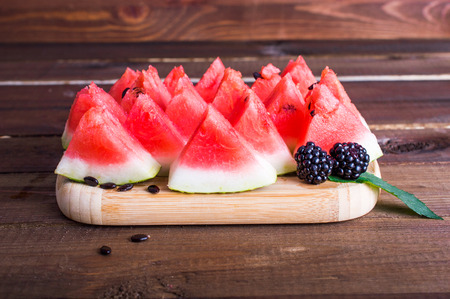 Triangular pieces of watermelon with seeds, blackberries and mint on a wooden board. On a wooden background.の写真素材