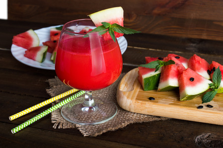 fresh watermelon in a glass with straws on a napkin. The triangular slices of watermelon on a plate with mint leaves and wooden board. On a wooden background.の写真素材