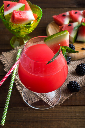 fresh watermelon in a glass with straws on a napkin. Triangular pieces of watermelon with mint and blackberries on a wooden board and a bowl. On a wooden background.の写真素材