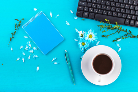Blue Notepad, blue pen, grass branch, blue chamomile flovers and petals, black keyboard, black coffee in a white cup and saucer on a blue background. Flat lay.の写真素材