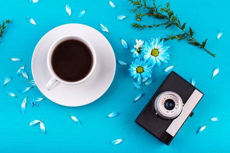 black and silver camera, grass branch, blue chamomile flovers and petals,  black coffee in a white cup and saucer on a blue background. Flat lay.の写真素材