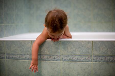 little cute baby bathing, hangs from the side of the bathtub and stretches down with one hand. against the background of a green bathroom in blur.の写真素材