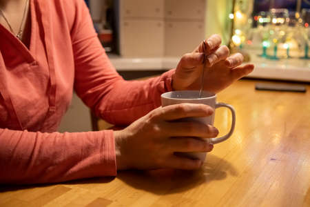 close-up female hands hold a white cup with a warm drink and mix with a teaspoon on a wooden table....