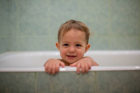 A cute Caucasian baby peeks out of the bathtub, put hands on the side of the bath and looks at the...