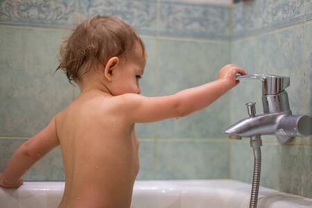 a little cute child, while bathing in the bath, plays with a water tap, turns it on and off. against the background of a green bathroom in blur close-up, soft focusの写真素材